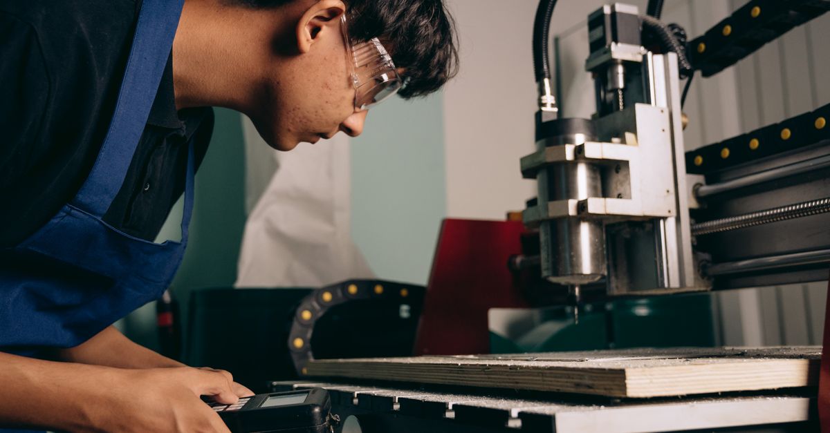 A focused young man wearing safety goggles operates a CNC machine in an industrial workshop.